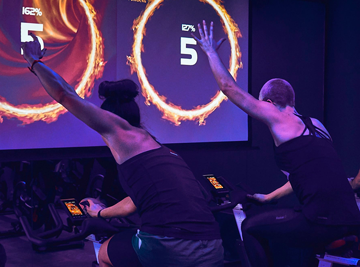Two participants are on spin bikes facing a big screen showing the number 5. They have their arms in the air quite evidently engrossed in the spin class. There is dramatic lighting which sets a energetic tone to the image