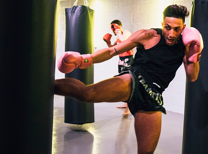 A male fighter is kicking a muay thai kick bag in the foreground of the image, wearing muay thai shorts and boxing gloves. In the background another boxer can be seen showing that they are partaking in a class